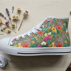 Puede incluir: Una zapatilla de ca&ntilde;a alta con un vibrante estampado floral tropical de hojas verdes, orqu&iacute;deas rosas y flores de hibisco amarillas. El calzado tiene una puntera de goma blanca, cordones blancos y una suela blanca con una fina raya negra lateral.