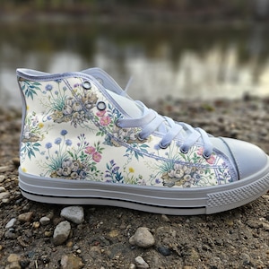 Puede incluir: Una zapatilla de caña alta con parte superior de lona color crema y un estampado botánico detallado de acianos azules, rosas rosadas y bulbos de ajo. El calzado incluye cordones gris claro, puntera de goma gris a juego y suela gris con raya negra.