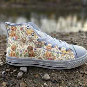 Könnte beinhalten: Ein High-Top-Sneaker mit einem cremefarbenen Canvas-Obermaterial, das mit einem sich wiederholenden Muster aus braunen und goldenen Doodle-Hunden sowie bunten Wildblumen und Grünpflanzen bedruckt ist. Der Schuh hat hellgraue Schnürsenkel, eine passende Zehenkappe und eine Gummisohle.