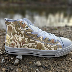 Puede incluir: Una zapatilla de caña alta con una parte superior de lona color crema con un estampado botánico detallado de flores de banksia marrones, follaje verde y serpientes entrelazadas. El calzado incluye cordones blancos, ojales de metal plateado.