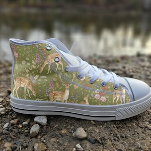 May include: A high-top trainer featuring a sage green canvas upper with a repeating pattern of deer, fawns, and various wildflowers. The shoe includes white laces, a white rubber toe cap, a white rubber sole with a thin black stripe, and silver eyelets.