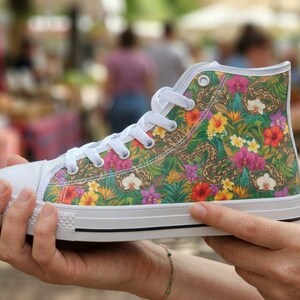 Puede incluir: Una zapatilla de ca&ntilde;a alta con un estampado tropical vibrante de hojas verdes, flores de hibisco amarillas y rojas, y orqu&iacute;deas moradas. El calzado tiene una puntera de goma blanca, cordones blancos y una suela blanca con una l&iacute;nea negra.