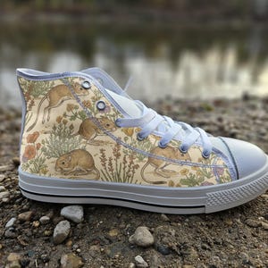 May include: A high-top sneaker featuring a cream-colored canvas upper with a repeating illustration of small brown rodents, orange poppies, and green foliage. The shoe includes white laces, a white rubber toe cap, and a white sole with black trim.