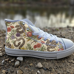 May include: A high-top trainer featuring a cream-coloured canvas upper with a repeating pattern of brown snakes, red flowers, and green foliage. The shoe includes white laces, a white rubber toe cap, and a white rubber sole with a thin black stripe.