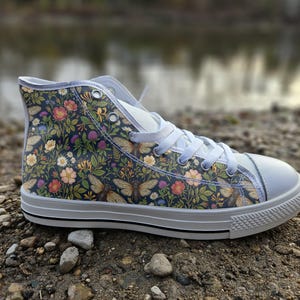Könnte beinhalten: Ein High-Top-Sneaker mit einem dunkelblauen Obermaterial aus Segeltuch, das mit einem sich wiederholenden Muster aus bunten Blumen, grünen Blättern und Zikaden bedruckt ist. Der Schuh hat weiße Schnürsenkel, eine weiße Gummikappe und eine weiße Sohle.