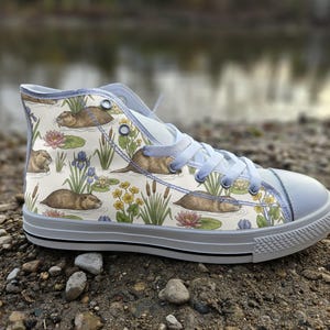 May include: A high-top trainer featuring a cream-coloured canvas upper with a repeating pattern of brown beavers, green cattails, yellow flowers, and pink water lilies. The shoe includes light blue laces, a white rubber toe cap, and a white sole.