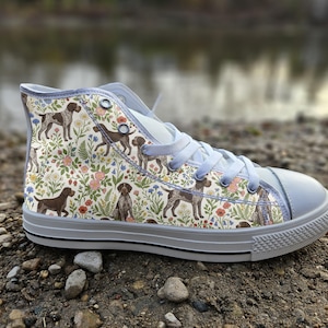 May include: A high-top sneaker featuring a cream-colored canvas upper with a repeating pattern of brown and white dogs interspersed with colorful wildflowers, green foliage, light blue laces, and a white rubber toe cap and sole.