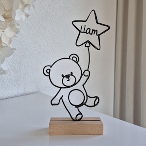 Könnte beinhalten: Ein schwarz-weißer Metallbär, der einen sternförmigen Ballon mit dem Namen "Liam" hält. Der Bär ist auf einem kleinen, rechteckigen Holzsockel montiert. Der Hintergrund umfasst eine weiße Vase mit weißen Blumen.