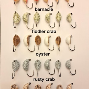 May include: A collection of fishing lures, each with a unique design and color scheme. The lures are labeled "ghost shrimp," "barnacle," "fiddler crab," "oyster," and "rusty crab." Each lure is attached to a silver hook.