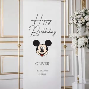 A banner with a Mickey Mouse face and the text "Happy Birthday Oliver" is displayed in a room with a floral arrangement in the background.
