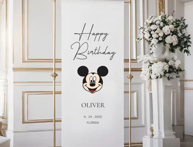 A banner with a Mickey Mouse face and the text "Happy Birthday Oliver" is displayed in a room with a floral arrangement in the background.
