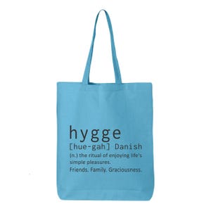 May include: A light blue tote bag with the word "hygge" printed in black. The definition of "hygge" is also printed on the bag, describing the Danish concept of enjoying life's simple pleasures, friends, family, and graciousness.
