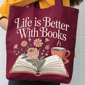 Puede incluir: Un bolso tote de color granate profundo con el texto blanco "Life is Better With Books" sobre una ilustración de un libro abierto con un ramo de flores rosas saliendo de las páginas y una taza de café naranja humeante a la derecha.