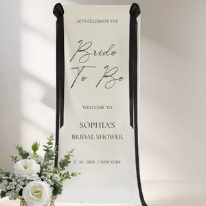 a banner that reads "Let's celebrate the Bride to Be" and "Welcome to Sophias Bridal shower 9. 24. 2020 / NEW YORK". There is also a vase of white flowers placed next to the banner.