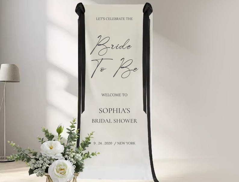 a banner that reads "Let's celebrate the Bride to Be" and "Welcome to Sophias Bridal shower 9. 24. 2020 / NEW YORK". There is also a vase of white flowers placed next to the banner.