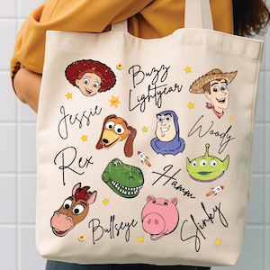 May include: A cream-colored canvas tote bag featuring colorful illustrations of Toy Story characters including Jessie, Buzz Lightyear, Woody, Rex, Hamm, Bullseye, Slinky, and an alien. The names of the characters are written in black script text.