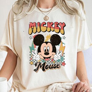 Könnte beinhalten: Cremefarbenes T-Shirt mit einem Retro-Mickey-Mouse-Motiv. Das Design zeigt die Worte "Mickey Mouse" in einer farbenfrohen Vintage-Schriftart, wobei Mickey das Friedenszeichen zeigt. Sterne und Blitze tragen zur verspielten Ästhetik bei.