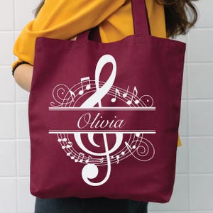 May include: Burgundy tote bag with a white musical note design. The design includes a treble clef, a bass clef, musical notes, and the name "Olivia" in a stylish font. The bag has a simple, practical design.