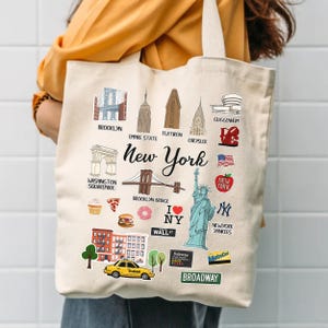 Könnte beinhalten: Cremefarbene Tragetasche mit Illustrationen von New Yorker Wahrzeichen, darunter die Brooklyn Bridge und die Freiheitsstatue. Der Text "New York" ist ebenfalls auf der Tasche zu sehen.