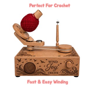 May include: A wooden yarn ball winder featuring a dark red yarn cake on a spindle, a belt-driven mechanism, and a control knob. The base is engraved with the text "WIND Joy CREATE," floral flourishes, birds, and a yarn ball graphic.