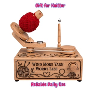 May include: A wooden yarn ball winder with a red yarn cake on the spindle. The base features engraved text reading "WIND MORE YARN WORRY LESS" with decorative floral and yarn ball motifs, a control knob, and a belt-driven mechanism.