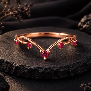 May include: A rose gold V-shaped ring featuring five round-cut vibrant pinkish-red gemstones set along the band. The metal has a polished finish, and the design follows a chevron silhouette with the stones spaced evenly across the front arc.