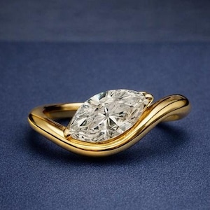 Può includere: Un anello con diamante taglio marquise e fascia in oro. Il diamante è incastonato in un design unico e fluido. L'anello è esposto su una scatola blu scuro, con una composizione floreale sfocata sullo sfondo. Un gioiello.
