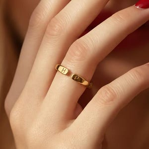 Könnte beinhalten: Ein goldener Bandring mit zwei kleinen, eingravierten Buchstaben H und P auf der Vorderseite. Der Ring hat eine glatte, polierte Oberfläche und ein schlichtes, abgerundetes Design. Das Metall wirkt wie Gelbgold und reflektiert das Licht.