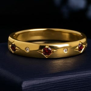 Può includere: Un anello d'oro con una serie di pietre rosse e piccole pietre trasparenti alternate. L'anello ha una finitura lucida ed è incastonato su uno sfondo blu scuro. Le pietre preziose sono incastonate a forma di stella.
