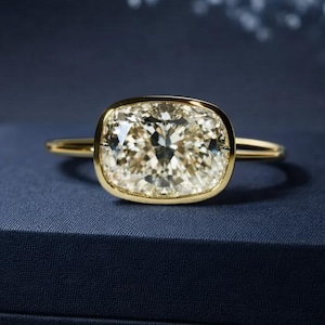 May include: A gold ring with a large, cushion-cut diamond. The diamond is set in a bezel setting and sits atop a gold band. The ring is displayed on a dark blue box, with a blurred background.