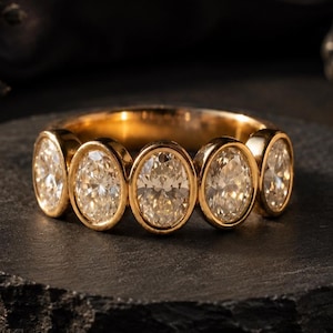 May include: A gold band ring featuring five oval-cut clear stones set in a bezel style. The stones are arranged horizontally across the front of the band, showcasing a polished metallic finish and faceted surfaces that reflect light.