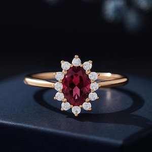 Op de afbeelding: Een gouden ring met een grote, ovaal geslepen, dieprode edelsteen, omringd door een halo van kleinere, ronde, witte edelstenen. De ring heeft een dunne, gepolijste gouden band.