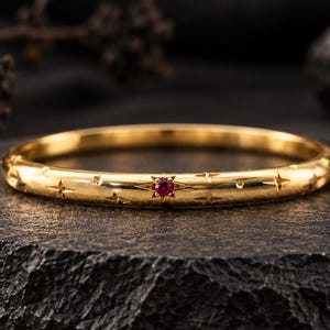 Op de afbeelding: Een gouden ring met een gepolijste afwerking, gegraveerde stermotieven en een enkele kleine, ronde, rode edelsteen in het midden. De ring heeft een glad, afgerond profiel en een consistente breedte over het gehele cirkelvormige ontwerp.