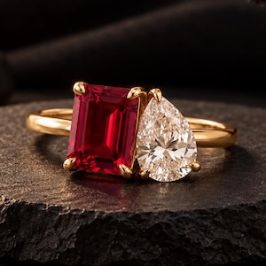 Può includere: Un anello in oro con una pietra preziosa rosso intenso dal taglio rettangolare a smeraldo posizionata accanto a una pietra bianca trasparente a forma di goccia. Le due pietre sono incastonate su un sottile anello in oro lucido.