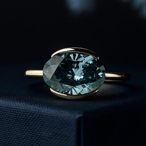 Può includere: Un anello d'oro con una grande pietra preziosa ovale color verde acqua. La pietra preziosa è incastonata in una lunetta e la fascia è in oro sottile e lucido. L'anello è esposto su una superficie blu scuro, con uno sfondo sfocato.
