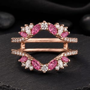 May include: A rose gold ring guard featuring two bands connected by a bridge. The design is adorned with marquise-cut pink stones and round white stones arranged in a floral-inspired pattern along the top and bottom edges of the ring structure.