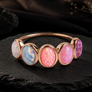 May include: A rose gold ring featuring five oval-shaped cabochon stones arranged in a row. The stones display a gradient of colors, transitioning from a milky white on the left to iridescent blue, vibrant pink, and a deeper magenta on the right.