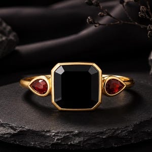 May include: A gold ring featuring a large, square-cut black stone in the center, flanked by two smaller, pear-shaped red stones on either side. The band is polished gold, and the setting holds the stones securely in a symmetrical, horizontal arrangement.