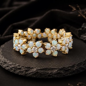 Può includere: Un anello color oro caratterizzato da una fascia continua di motivi floreali interconnessi. Ogni fiore è realizzato con sei petali rotondi in opale bianco iridescente che circondano una singola pietra rotonda, chiara e brillante al centro.