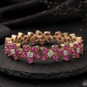 May include: A gold-toned bracelet featuring a repeating floral pattern. Each flower is composed of five vibrant pink circular stones surrounding a single clear white center stone. The metal links connect the floral segments into a circular band.