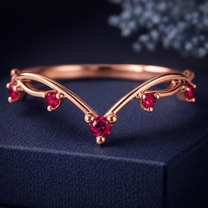 May include: A rose gold V-shaped ring featuring five round-cut vibrant red rubies set along the band. The metal has a polished finish, and the design follows a chevron silhouette with the stones spaced evenly across the front of the band.