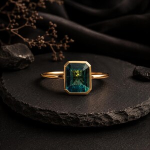 May include: A gold ring featuring a rectangular emerald-cut teal gemstone set in a smooth bezel setting. The band is thin and polished, showcasing a vibrant blue-green stone with visible facets that catch the light, centered on a dark surface.
