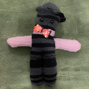 Puede incluir: Una muñeca hecha a mano con un cuerpo a rayas grises y negras, brazos rosas y un sombrero gris. La muñeca tiene ojos de botón negros y una pajarita con lentejuelas. La muñeca está hecha de calcetines y otros materiales artesanales.