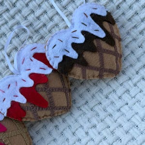May include: Felt heart-shaped ornaments resembling ice cream cones. The cones are brown with a waffle pattern, topped with white felt "ice cream" and red or brown "syrup". Each ornament has a white ribbon for hanging.