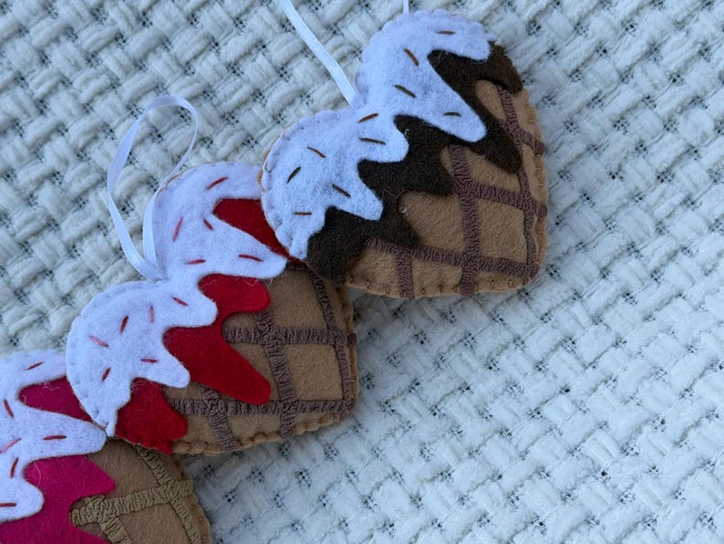 May include: Felt heart-shaped ornaments resembling ice cream cones. The cones are brown with a waffle pattern, topped with white felt "ice cream" and red or brown "syrup". Each ornament has a white ribbon for hanging.