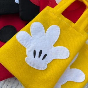 Może przedstawiać: Ż&oacute;łte filcowe torby z aplikacjami białych rękawic Myszki Miki. Torby mają pasujące ż&oacute;łte uchwyty i są zszyte białą nicią. W tle widoczne są inne torby w kolorze czerwonym i czarnym.