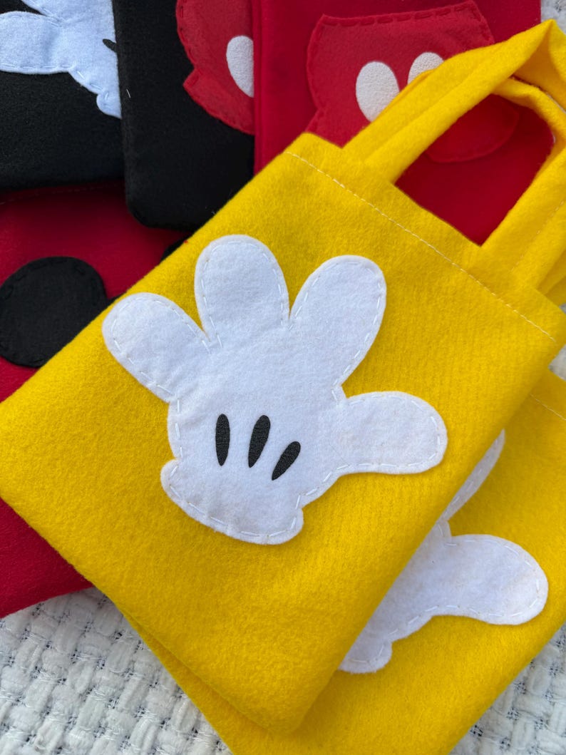 Może przedstawiać: Ż&oacute;łte filcowe torby z aplikacjami białych rękawic Myszki Miki. Torby mają pasujące ż&oacute;łte uchwyty i są zszyte białą nicią. W tle widoczne są inne torby w kolorze czerwonym i czarnym.