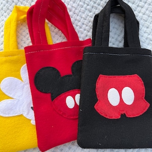 Może przedstawiać: Trzy małe filcowe torby w kolorze ż&oacute;łtym, czerwonym i czarnym. Ż&oacute;łta torba ma biały kwiat, czerwona torba ma twarz Myszki Miki, a czarna torba ma spodenki Myszki Miki. Każda torba ma pasujący uchwyt.