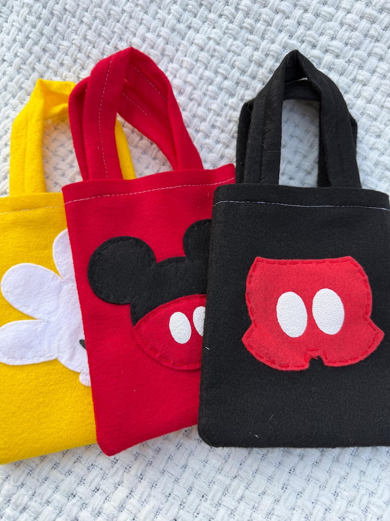 Może przedstawiać: Trzy małe filcowe torby w kolorze ż&oacute;łtym, czerwonym i czarnym. Ż&oacute;łta torba ma biały kwiat, czerwona torba ma twarz Myszki Miki, a czarna torba ma spodenki Myszki Miki. Każda torba ma pasujący uchwyt.