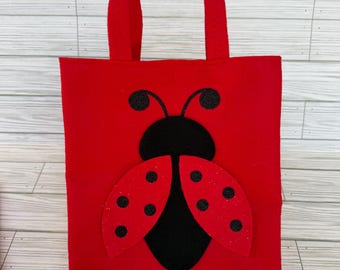 Può includere: Una borsa tote in feltro rosso brillante con un'applicazione a forma di coccinella sul davanti. La coccinella ha una testa nera con antenne e due ali rosse decorate con puntini neri glitterati. Include due manici in feltro rosso.